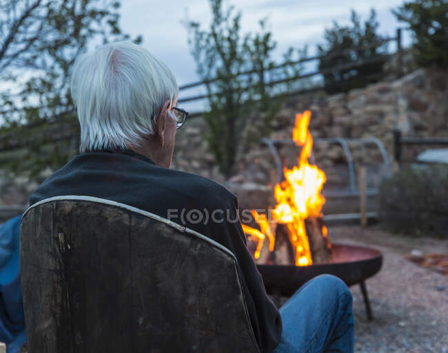 Visão traseira de um homem idoso sentado olhando registros queimando em uma fogueira — Fotografia de Stock