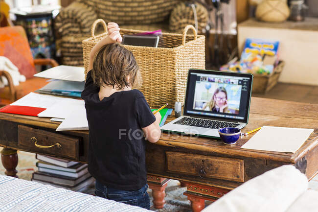 Ребенок работает дома, смотрит на учителя на экране онлайн уроки, поднимая руку, чтобы ответить на вопросы — стоковое фото