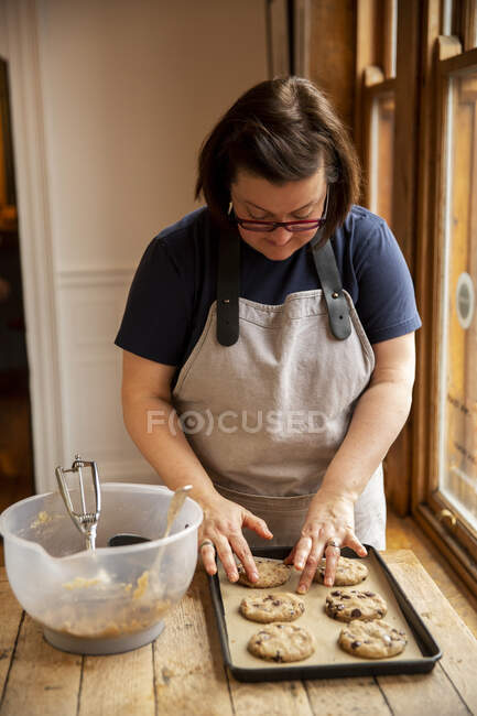 Donna che indossa occhiali e grembiule in piedi al tavolo di legno, cottura biscotti con gocce di cioccolato. — Foto stock