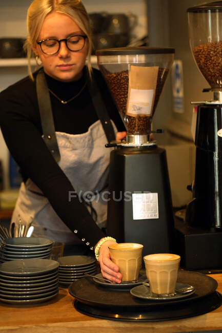 Mulher loira usando óculos e avental de pé no balcão em um café, colocando dois cafés lattes em uma bandeja. — Fotografia de Stock