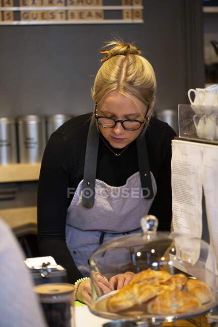 Blonde Frau mit Brille und Schürze steht in einem Café an der Espressomaschine und schreibt einen Zettel. — Stockfoto