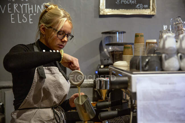 Blond woman wearing glasses and apron standing at espresso machine in a cafe, pouring milk into metal jug. — Stock Photo