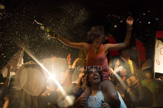 Revellers en un concierto al aire libre, un hombre sonriente cargando a una mujer sobre sus hombros, con los brazos extendidos, sosteniendo una botella de cerveza. - foto de stock