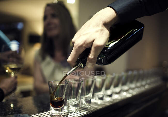 Primer plano del camarero vertiendo bebidas de la botella en una fila de vasos de chupito de pie en el mostrador de la barra, mujer sentada en el fondo. - foto de stock