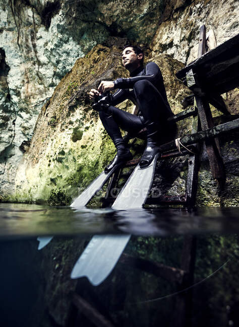 Mann in Neoprenanzug und Schwimmflossen sitzt auf einem Podest, Felsen im Hintergrund. — Stockfoto