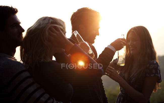Zwei Männer und zwei Frauen stehen bei Sonnenuntergang im Freien, halten Bierflaschen in der Hand und lächeln. — Stockfoto