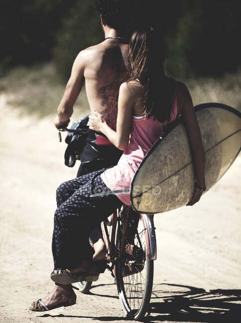Rear view of man and woman on a bicycle, woman sitting on rack, holding a surfboard. — Stock Photo