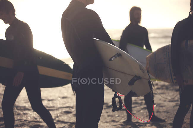 Quatro homens vestindo roupas de mergulho em pé em uma praia de areia, levando pranchas de surf. — Fotografia de Stock