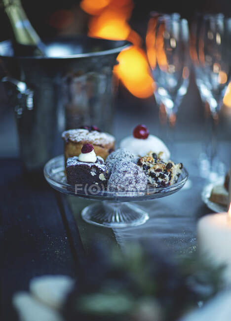 Close up of glass cake stand with a selection of cakes, champagne glasses and wine cooler in the background. — Fotografia de Stock