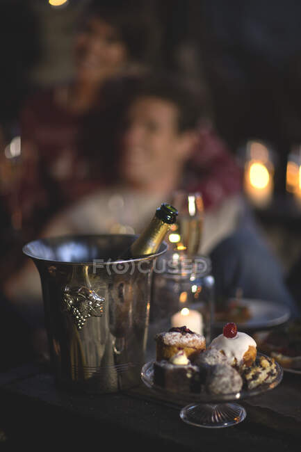 Primer plano de botella de champán en enfriador de vino de metal, soporte de pastel de vidrio con selección de pasteles, persona en el fondo. - foto de stock
