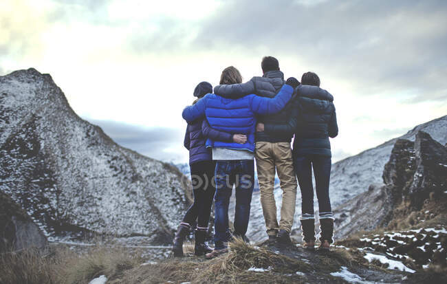 Вид сзади на четыре человека, стоящих рука об руку на горе, снежные вершины вдалеке. — стоковое фото