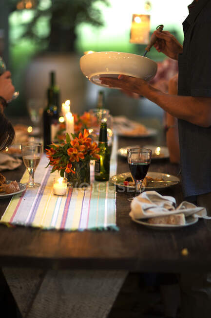Person, die an einem Tisch steht und eine Schale, Weingläser, Teller, Blumen und Kerzen auf dem Tisch hält. — Stockfoto