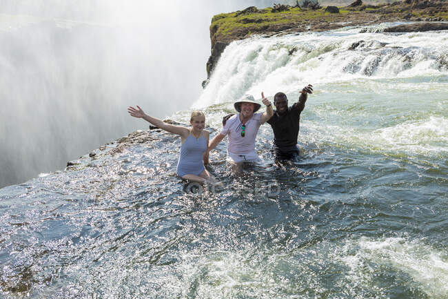 Three people, two men and young teenager standing in the waters of the Devil's Pool on the edge of the Victoria Falls, arms outstretched. — Stockfoto
