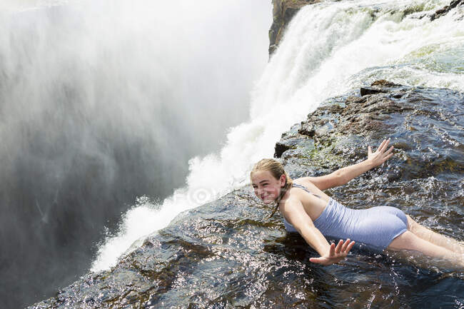 Chica joven en el agua en la piscina de los demonios acostada en su frente, los brazos extendidos, en el borde del acantilado de Victoria Falls. - foto de stock