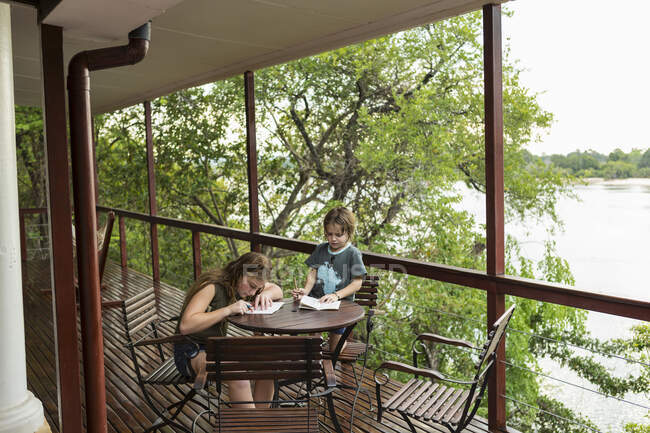 Two children drawing together at a table on a terrace overlooking a wide river. — Stock Photo