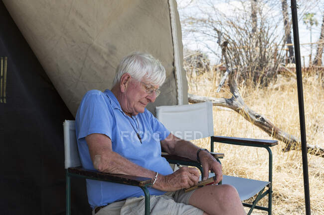 Senior man writing a journal seated outside a tent in a wildlife safari camp. — Stock Photo