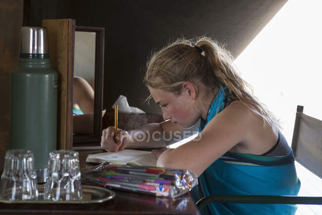 A twelve year old girl seated at a desk in a tent at a wildlife reserve camp, drawing. — Stock Photo