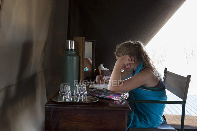 Una bambina di dodici anni seduta ad una scrivania in una tenda in un campo di riserva naturale, disegnando. — Foto stock