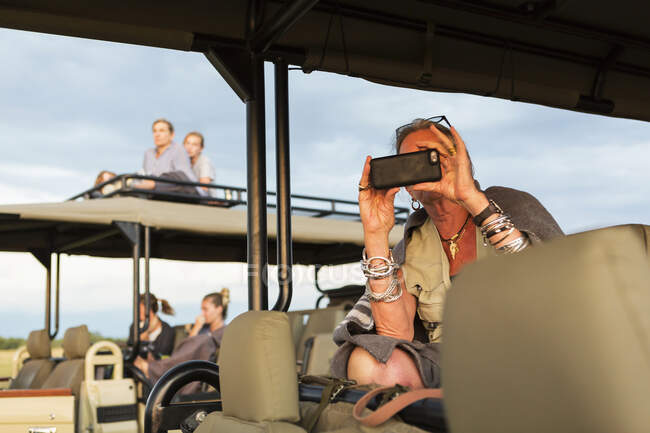 Mujer mayor tomando fotos con un teléfono inteligente desde un jeep safari. - foto de stock