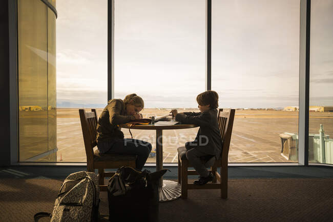 Un niño y su hermana mayor sentados en una mesa en un salón del aeropuerto, escribiendo y dibujando. - foto de stock