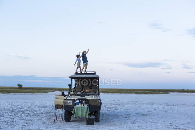 Hermano y hermana de pie en la parte superior de un vehículo de safari al atardecer en un paisaje de sal pan - foto de stock