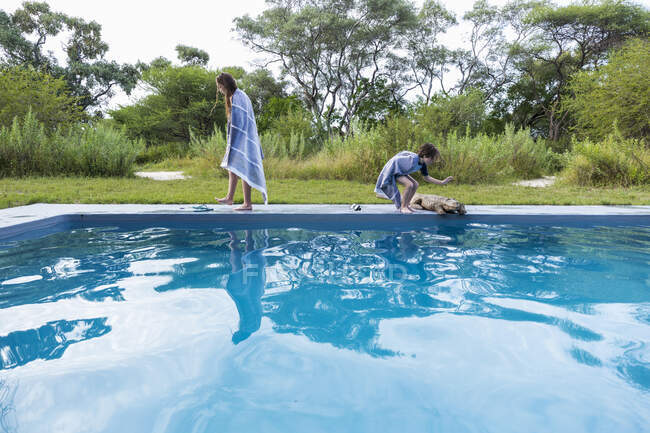 Two children by a swimming pool, one petting a large wooden crocodile at a resort. — Stock Photo