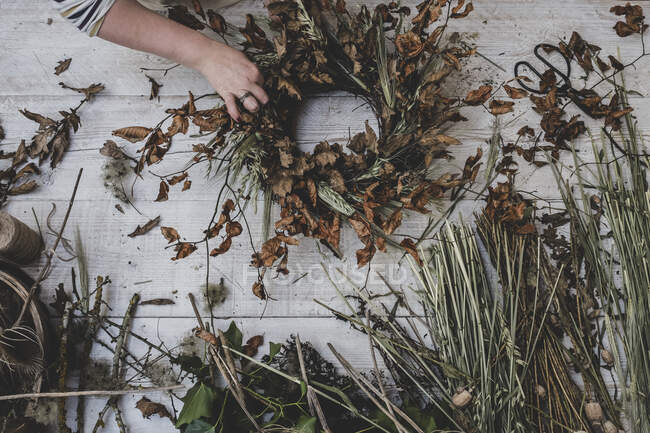 Mujer haciendo una pequeña corona de invierno de plantas secas, hojas marrones y ramitas, y cabezas de semillero. - foto de stock