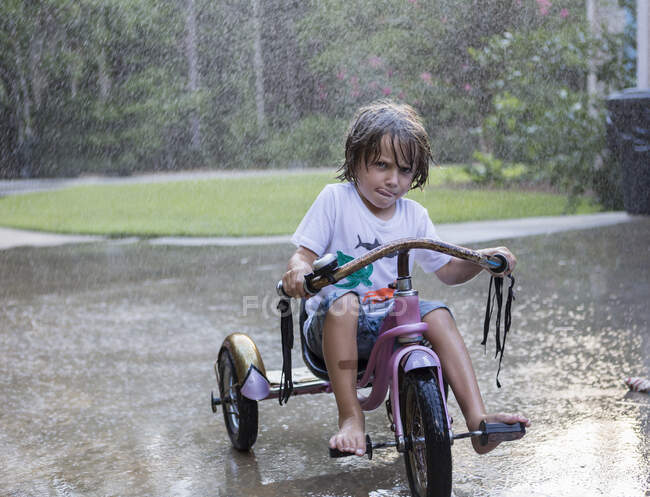 Niño de 5 años montando su bicicleta de triciclo bajo la lluvia - foto de stock