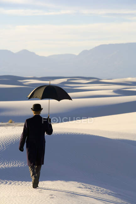 Homme vêtu d'un manteau et d'un costume noirs, chapeau et parapluie melon, dans un désert blanc de sable blanc. — Photo de stock