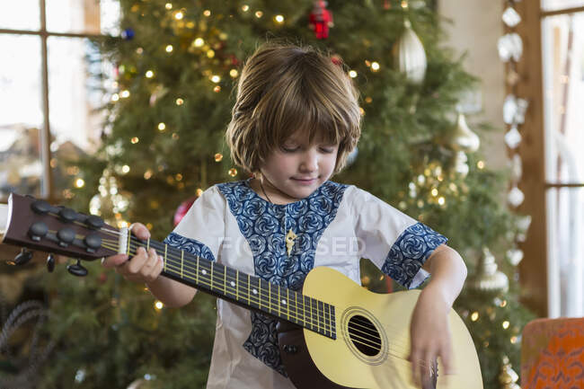 Улыбающийся 4-летний мальчик играет на гитаре на фоне рождественской елки — стоковое фото