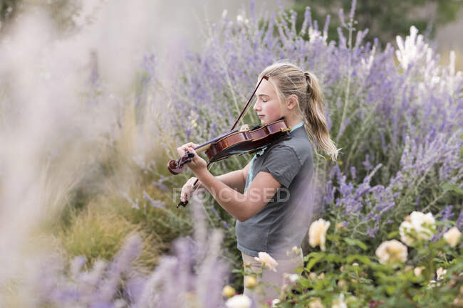 Adolescente de pé entre rosas floridas e arbustos, tocando um violino — Fotografia de Stock