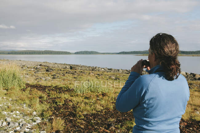 Жінка середнього віку, використовуючи бінокль, дивиться на далеке узбережжя затоки на узбережжі Аляски.. — стокове фото