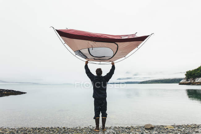 Чоловік, що тримає табір над головою, стоїть на скелястому пляжі, вхід на узбережжі Аляски.. — стокове фото