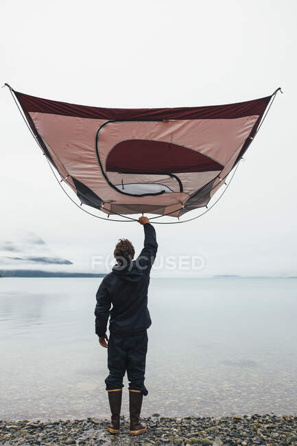 Uomo che tiene tenda da campeggio sopra la testa, in piedi sulla spiaggia rocciosa, una insenatura sulla costa dell'Alaska. — Foto stock