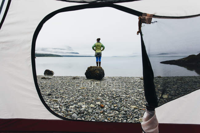 Vista a través de tienda de campaña puerta de la mujer de pie en la playa, una ensenada en la costa de Alaska. - foto de stock