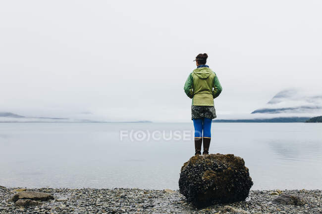 Mulher em pé em uma praia com vista para uma entrada costeira, névoa e nuvem baixa. — Fotografia de Stock