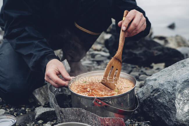 Un campista salvaje revolviendo comida caliente en una olla cocinando sobre un fuego. - foto de stock