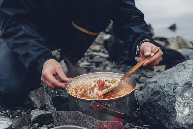 Un campista salvaje revolviendo comida caliente en una olla cocinando sobre un fuego. - foto de stock