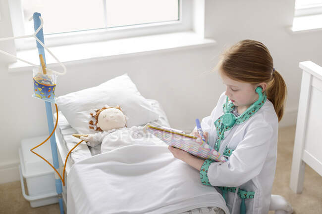 Giovane ragazza vestita da medico che scrive appunti accanto a fingere paziente nel letto d'ospedale finto — Foto stock