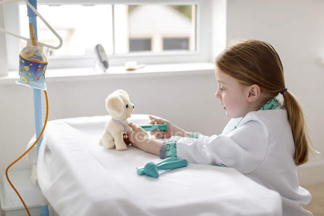 Молодая девушка, одетая как доктор, притворяющаяся, что лечит игрушечную собаку в больничной койке — стоковое фото