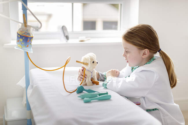 Chica joven vestida como médico fingiendo tratar perro de juguete en cama de hospital make-bleieve - foto de stock