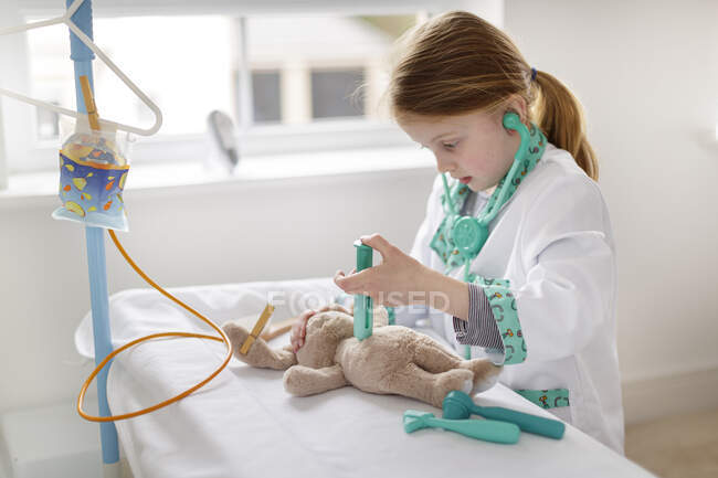 Jeune fille habillée en médecin prétendant traiter peluche jouet dans un lit d'hôpital imaginaire — Photo de stock