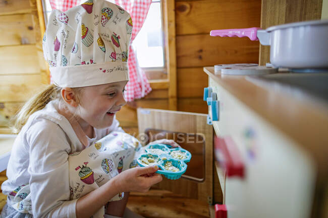Young girl in wendy house putting cup cakes in oven pretending to cook in kitchen — Stock Photo