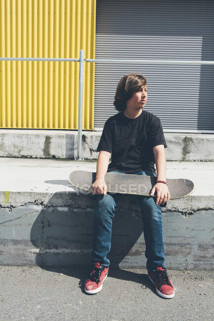 Adolescente posando sentado con monopatín frente a almacén urbano - foto de stock