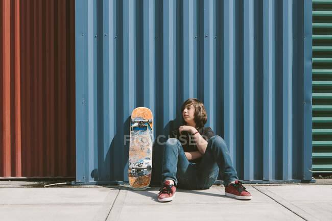 Teenage boy sitting with skateboard against warehouse wall — Stock Photo
