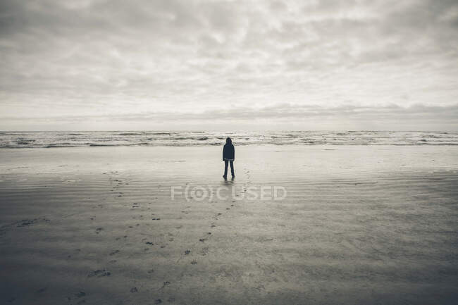 Teenage boy standing on vast beach, waves and overcast sky in distance — Stock Photo