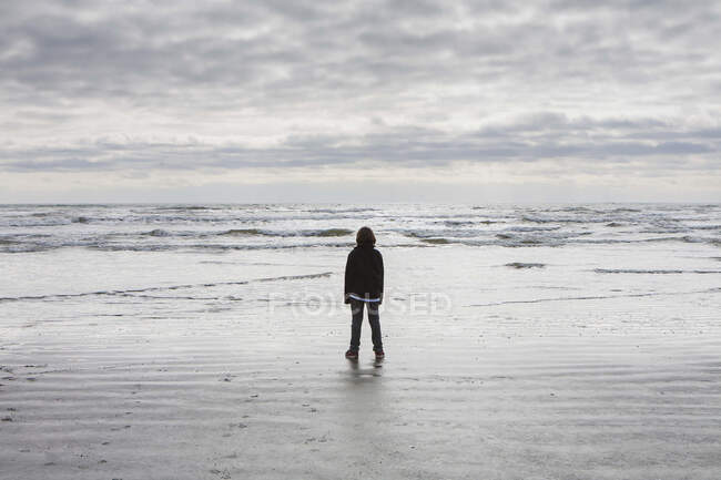 Teenage boy standing on vast beach, waves and overcast sky in distance — Stock Photo
