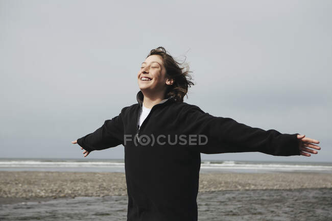 Teenage boy on beach with arms outstretched towards breeze, ocean in distance — Stock Photo