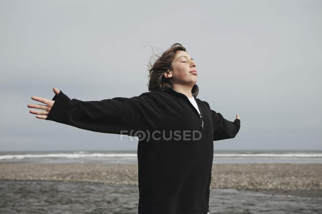 Мальчик-подросток на пляже с протянутыми к ветру руками, вдалеке океан — стоковое фото