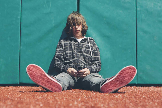 Хлопчик-підліток, що сидить на спортивному полі, тримає мобільний телефон — стокове фото